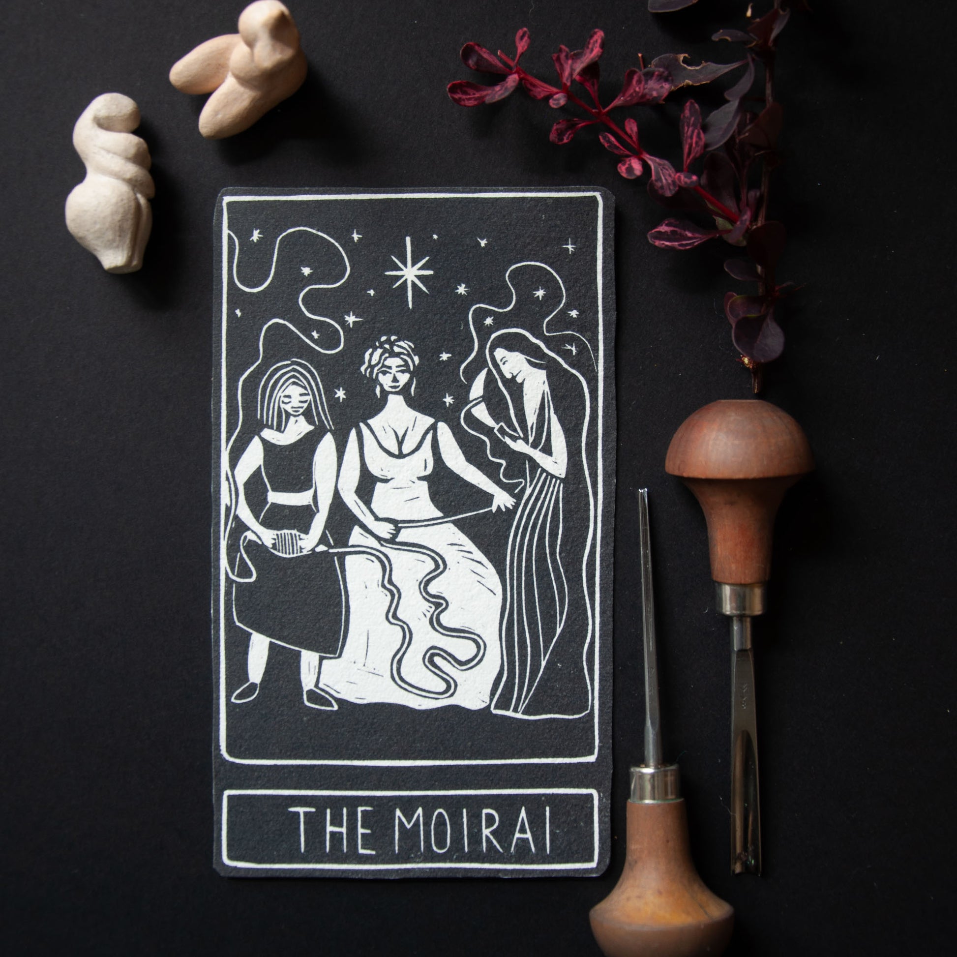 Tarot card titled 'The Moirai' with decorative items on a dark background