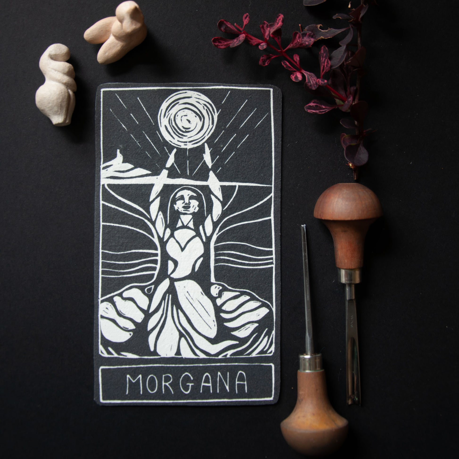 Tarot card featuring a woman with a moon symbol, surrounded by tools and natural elements on a dark background.