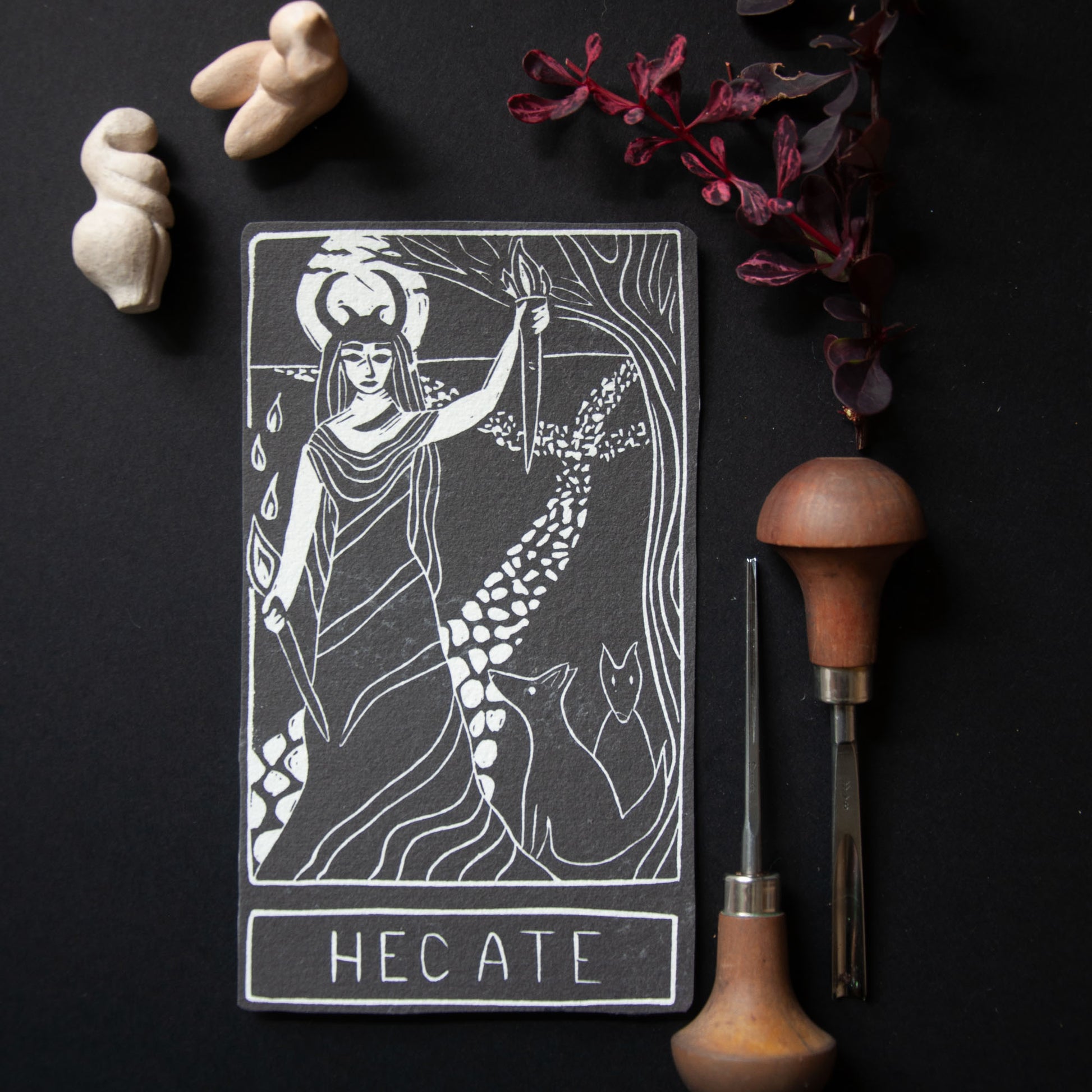 Tarot card featuring Hecate with tools and decor on a dark background