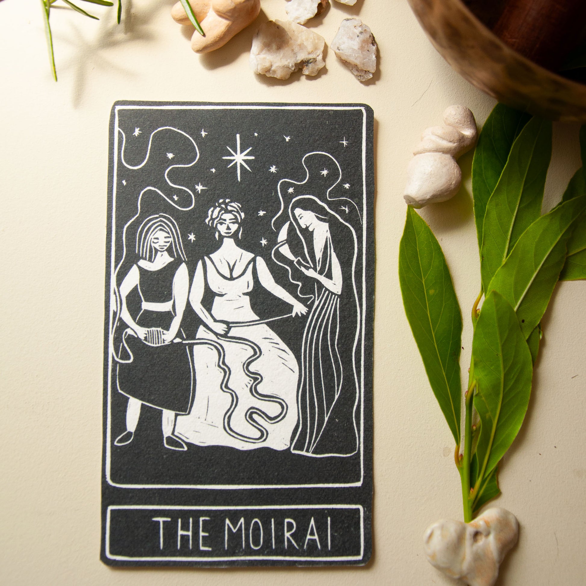 Tarot card titled 'The Moirai' with three women on a beige surface with leaves and stones.