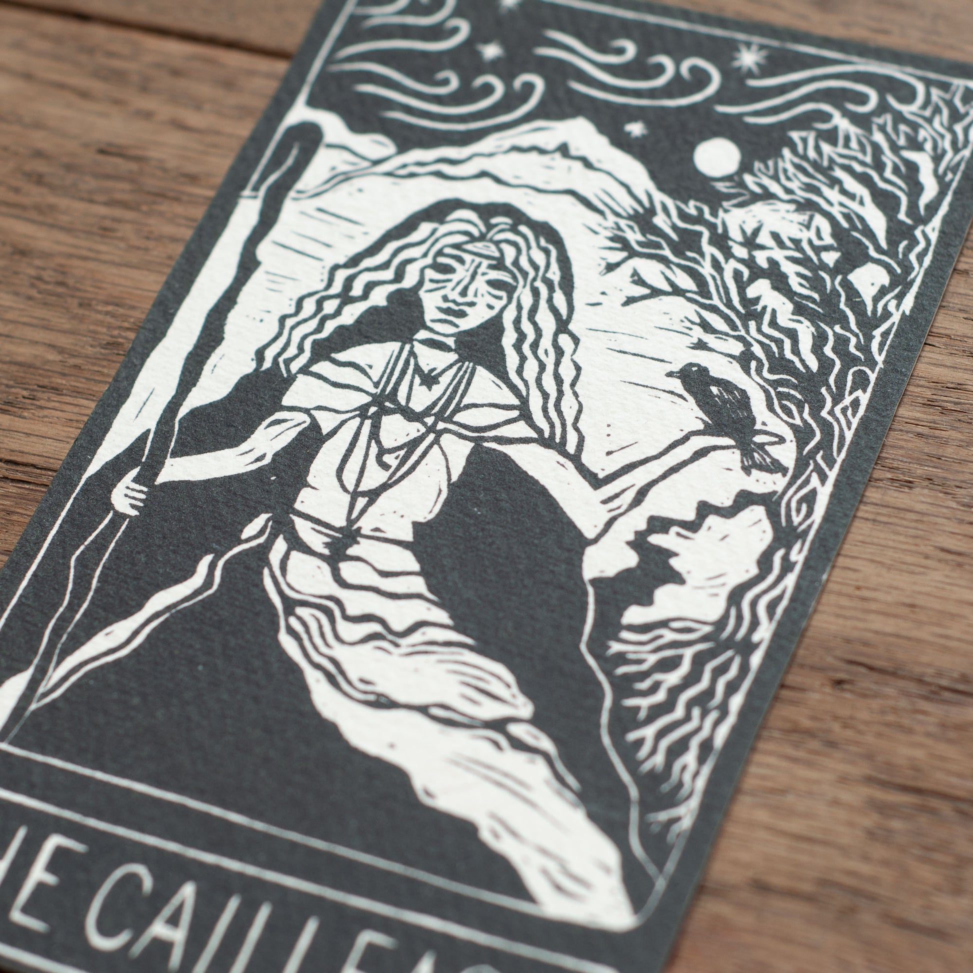Black and white illustration of a woman with a staff on a textured card, placed on a wooden surface.