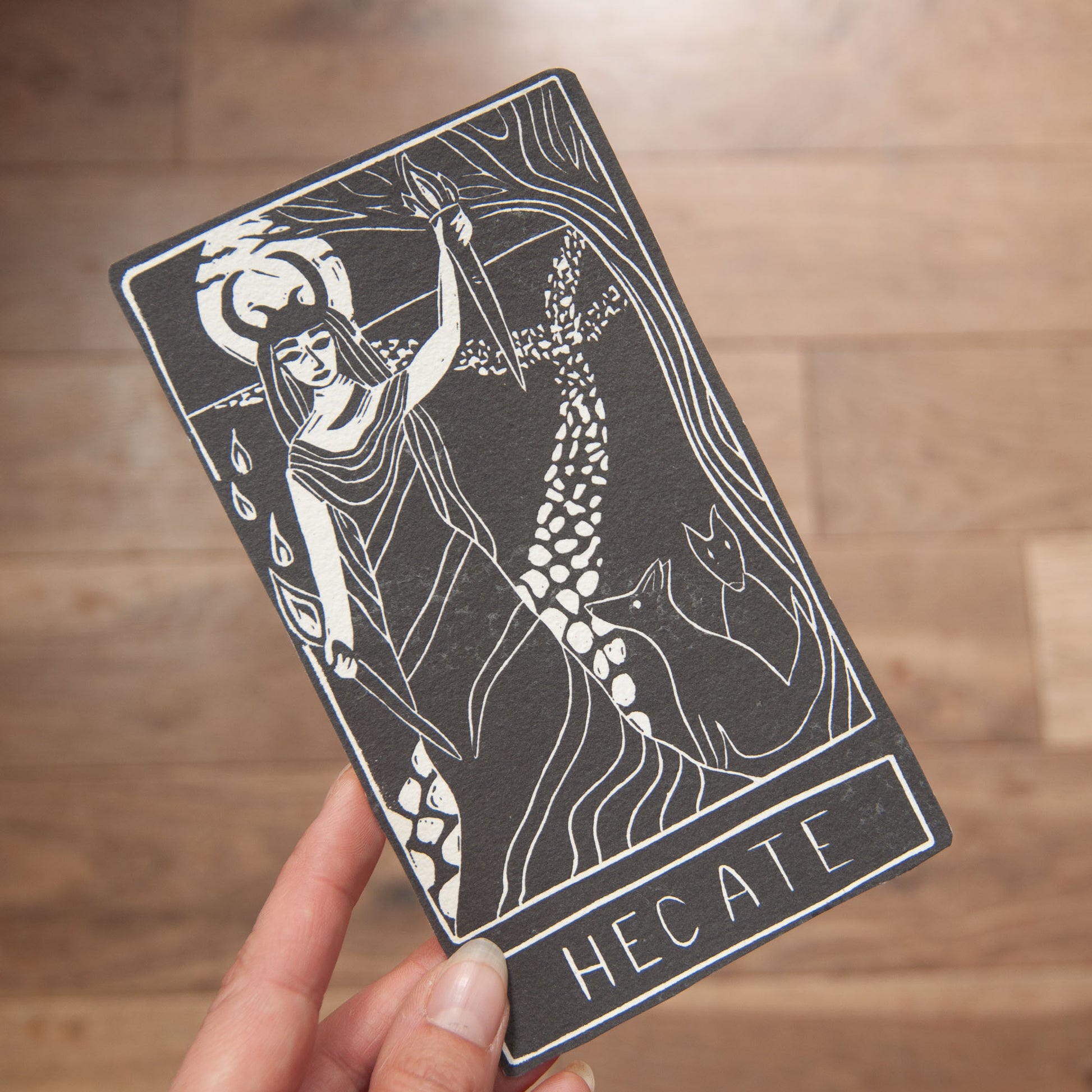 Card featuring an illustration of Hecate with a person holding it against a wooden background