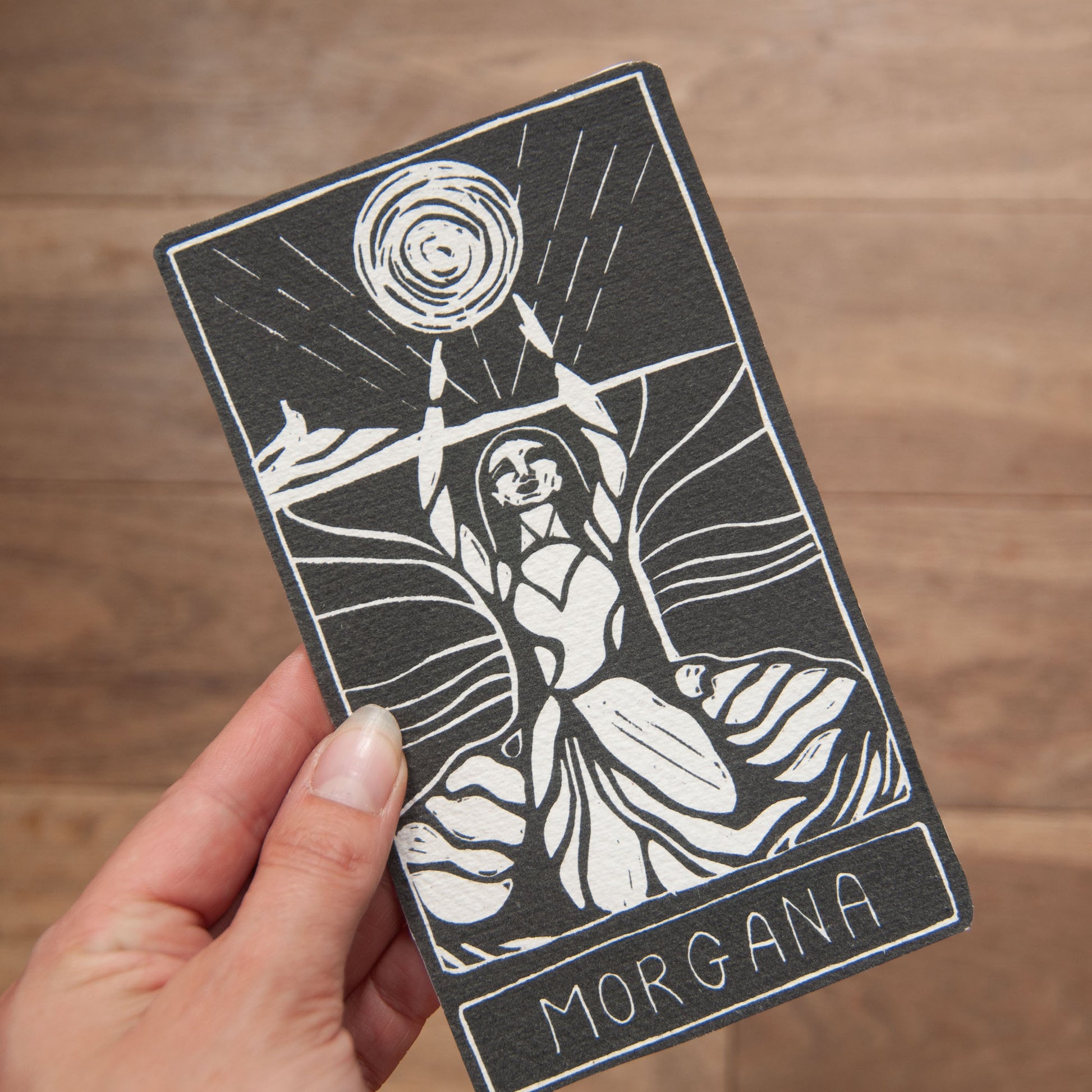 Tarot card featuring a black and white illustration of a figure holding a disc, with 'Morgana' written below, held by a hand on a wooden surface.