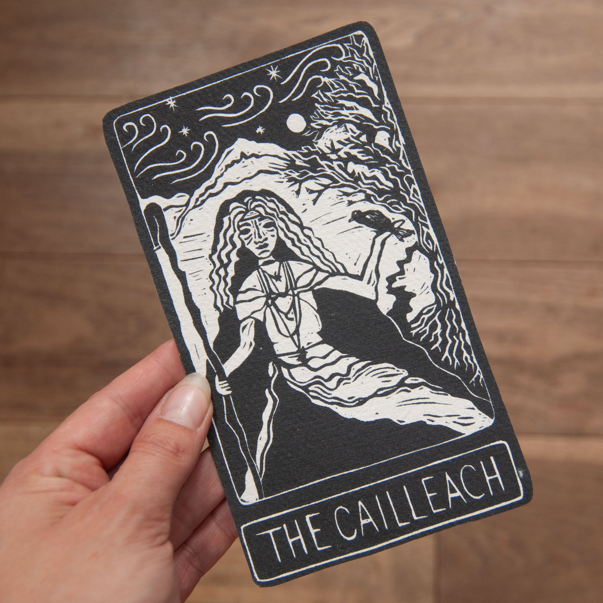 Card featuring 'The Cailleach' design held by a hand on a wooden surface