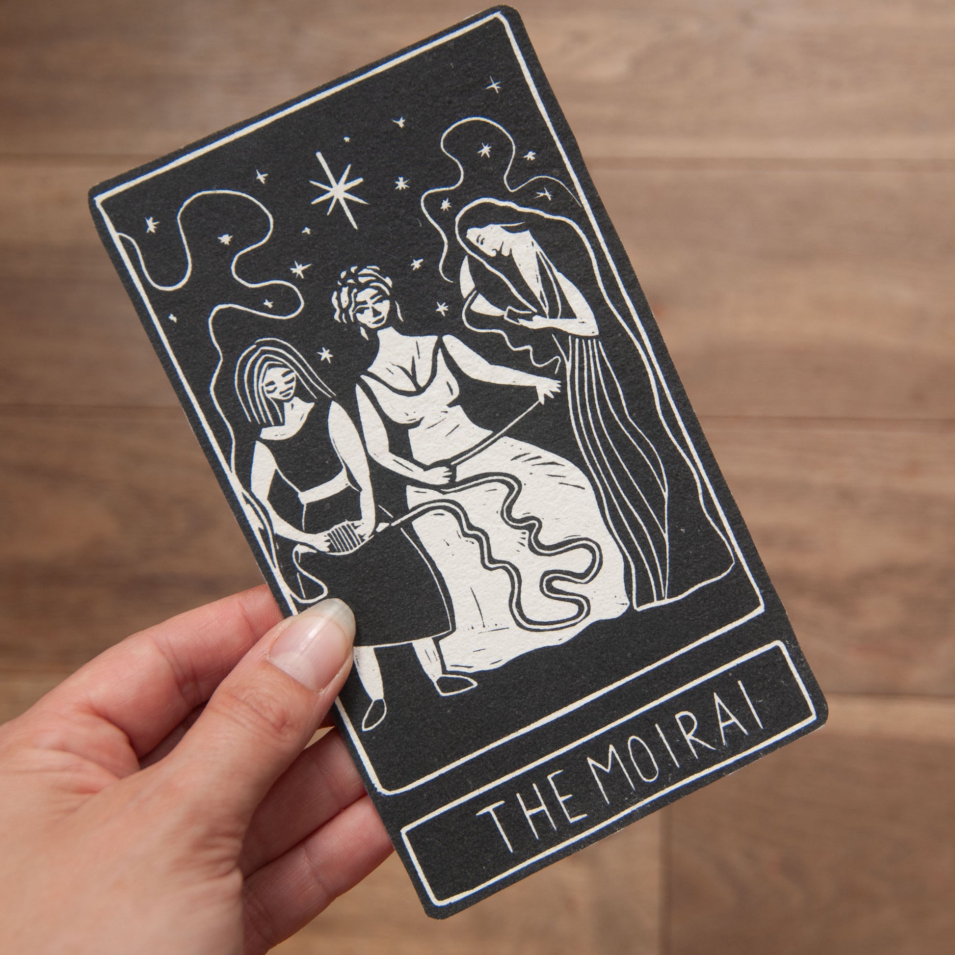 Hand holding a tarot card with 'The Moirai' design on a wooden surface