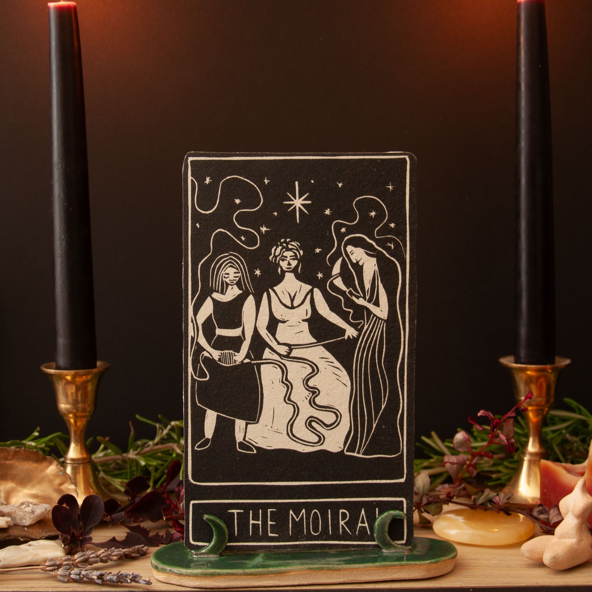Tarot card titled 'The Moira' with three women on a dark background with candles and herbs.