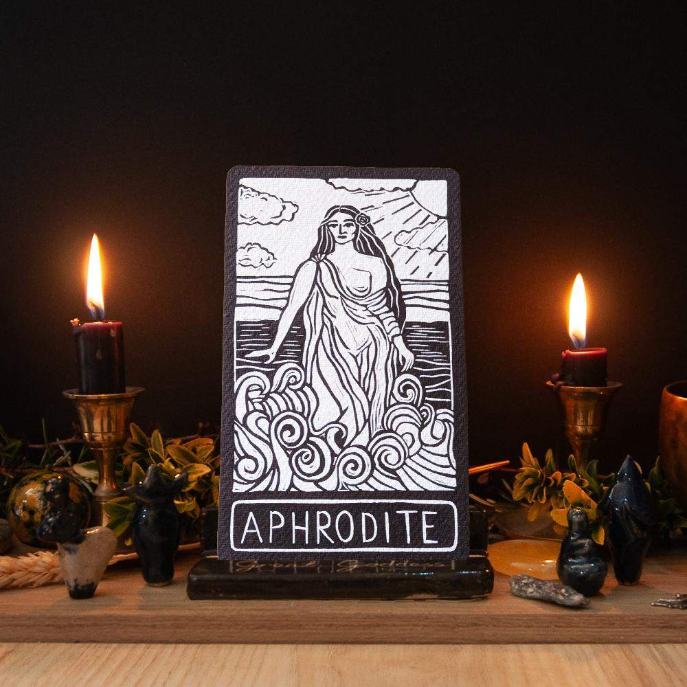APHRODITE ALTAR CARD with a black-and-white linocut-style goddess in flowing robes above swirling waves, with bold lettering