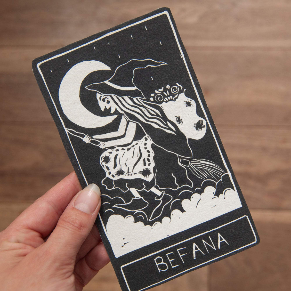 BEFANA ORACLE CARD in cream and black, with a vintage linocut witch riding a broom across clouds beneath a crescent moon.