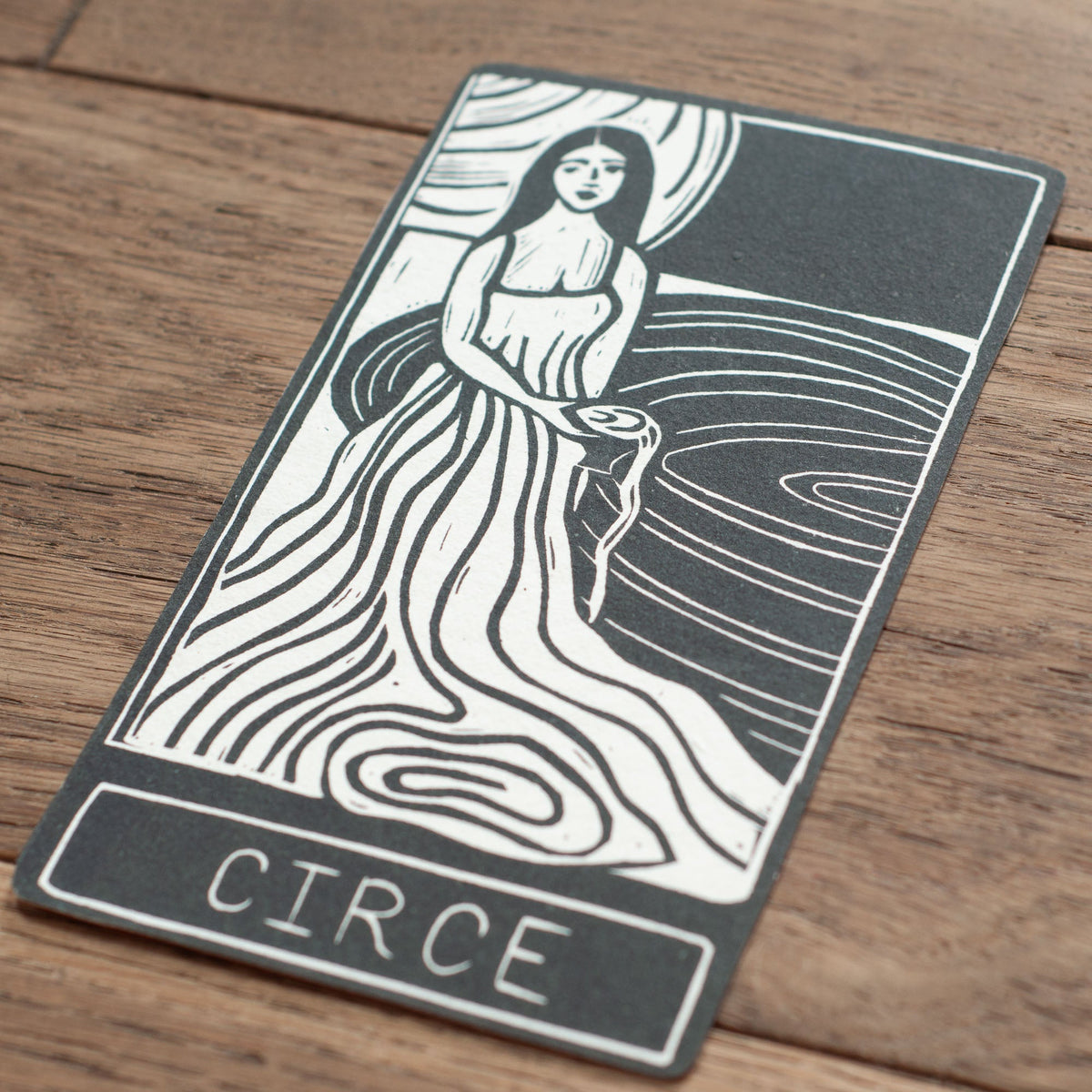 CIRCE ORACLE CARD in dark grey and cream, featuring a handmade linocut of a robed goddess beside flowing waves