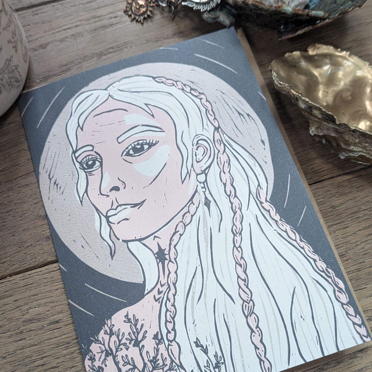 Frigg Greeting card featuring a pale blue and pink linocut portrait of a braided-haired goddess on charcoal cardstock.