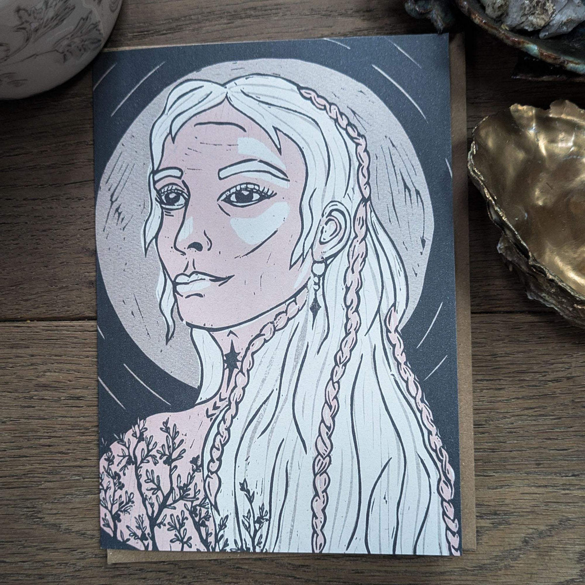 Frigg Greeting card with a pale blue and pink linocut portrait of a braided-haired Norse goddess on dark cardstock.