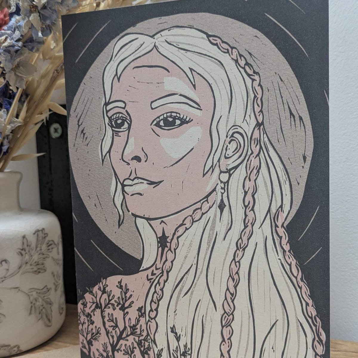 Frigg Greeting card with a pale pink and cream linocut portrait of a braided-haired goddess on dark charcoal cardstock.