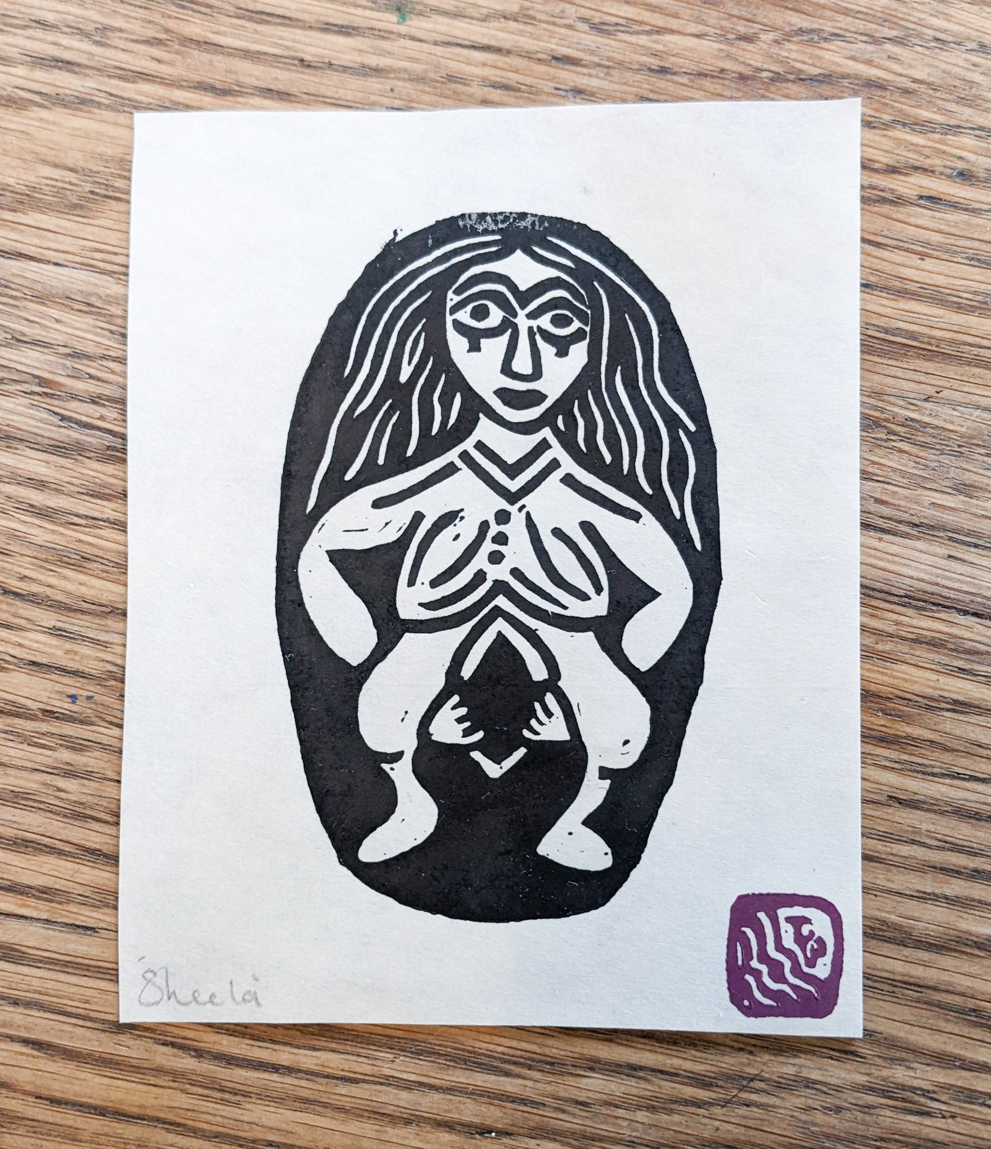 image of sheela na gig print, black ink on white delicate paper on a wooden background
