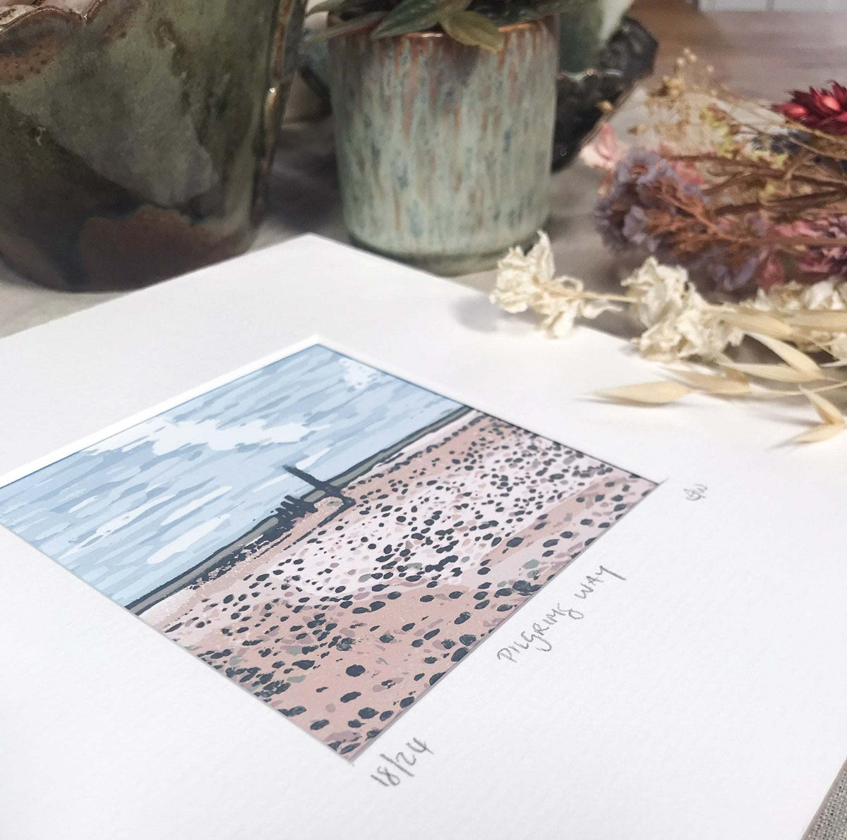 PILGRIM'S WAY linocut print in muted blue and earthy pink, depicting a coastal path through textured landscape.