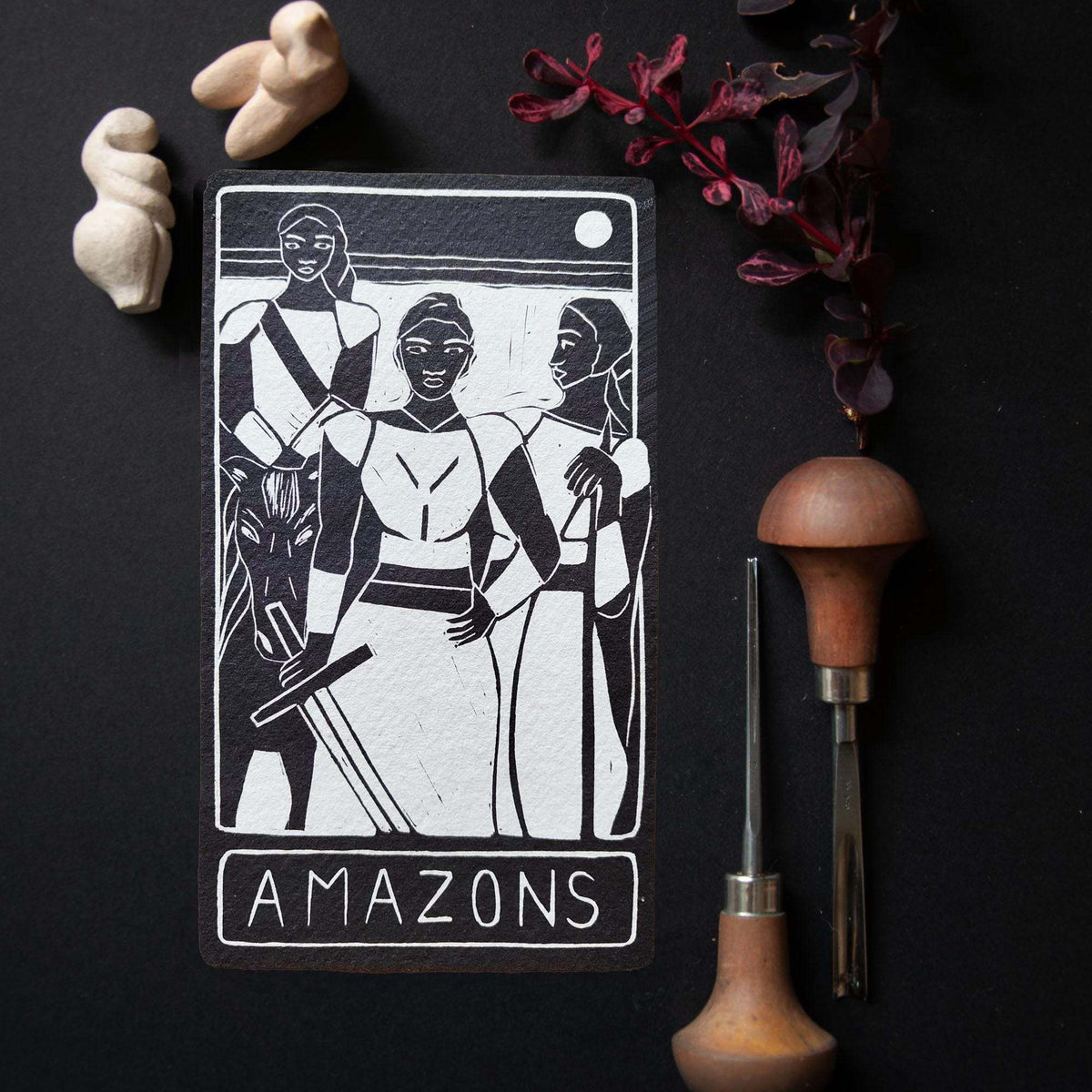THE AMAZONS ALTAR CARD with a black-and-white linocut of three warrior women, framed by a bold border and title