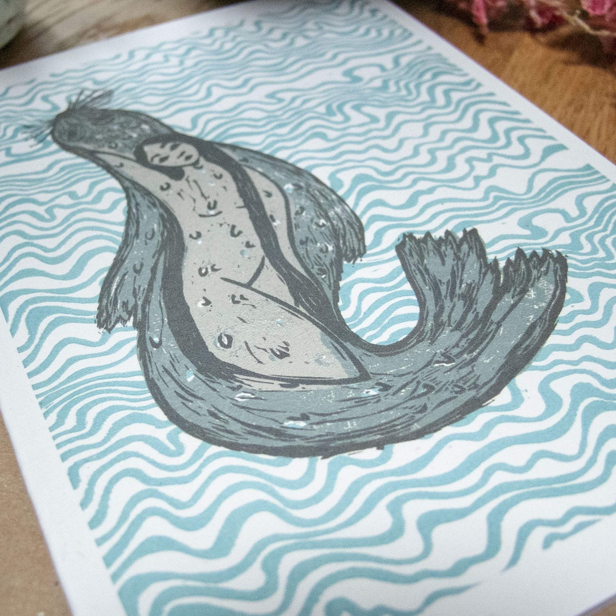 THE SELKIE Greeting card in natural cardstock, with a grey linocut seal-woman surrounded by teal wavy lines.