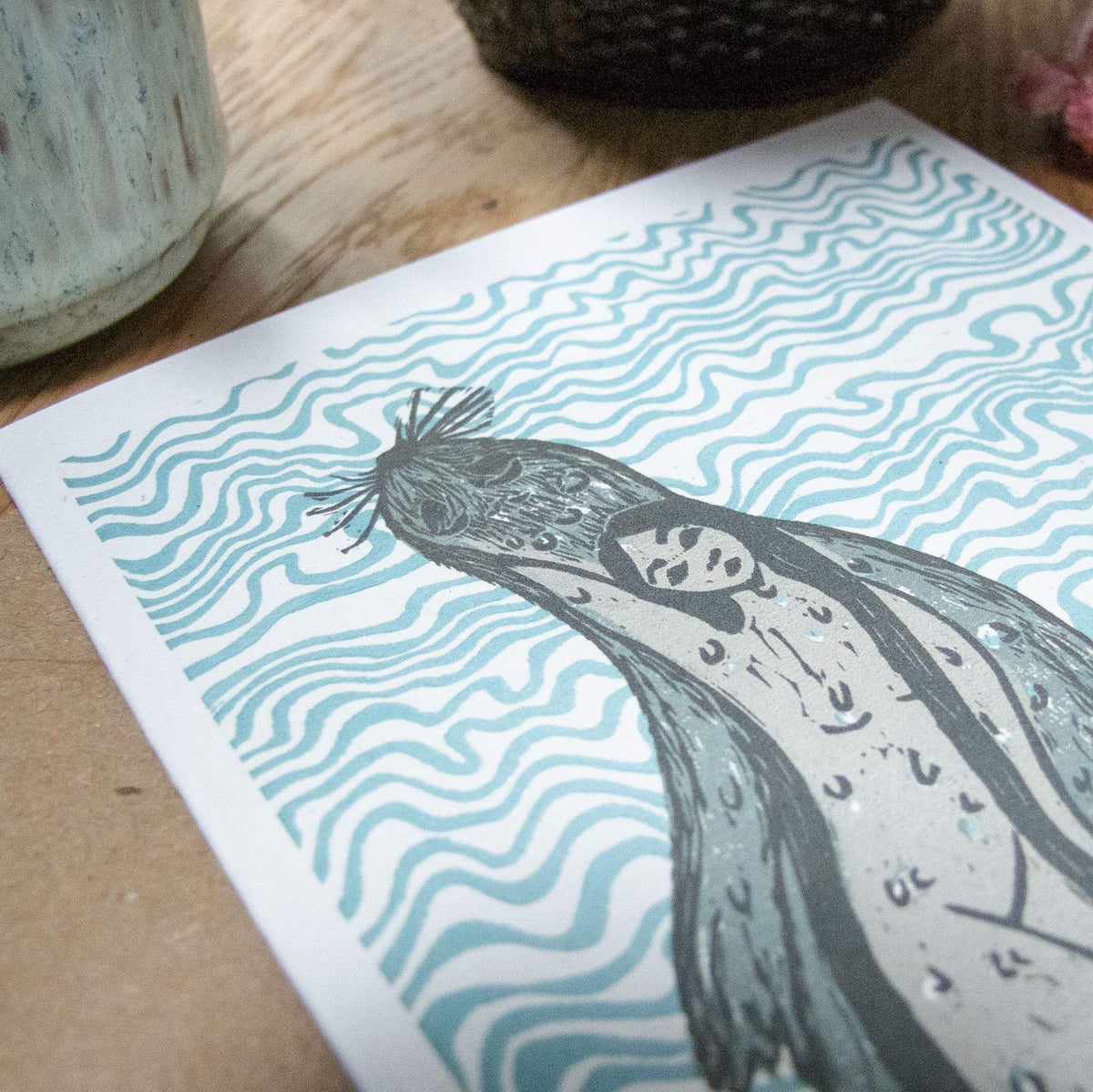 THE SELKIE Greeting card featuring a grey linocut selkie with a human face amid teal wavy lines on white cardstock.