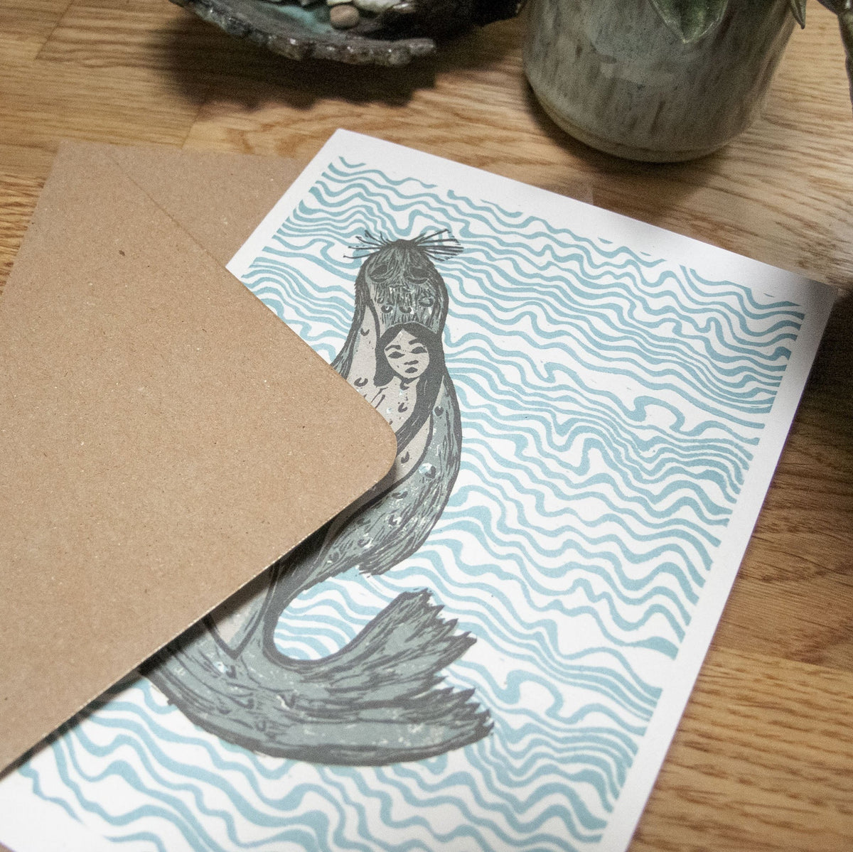 THE SELKIE Greeting card with a black linocut selkie and pale blue wavy water pattern on white cardstock.