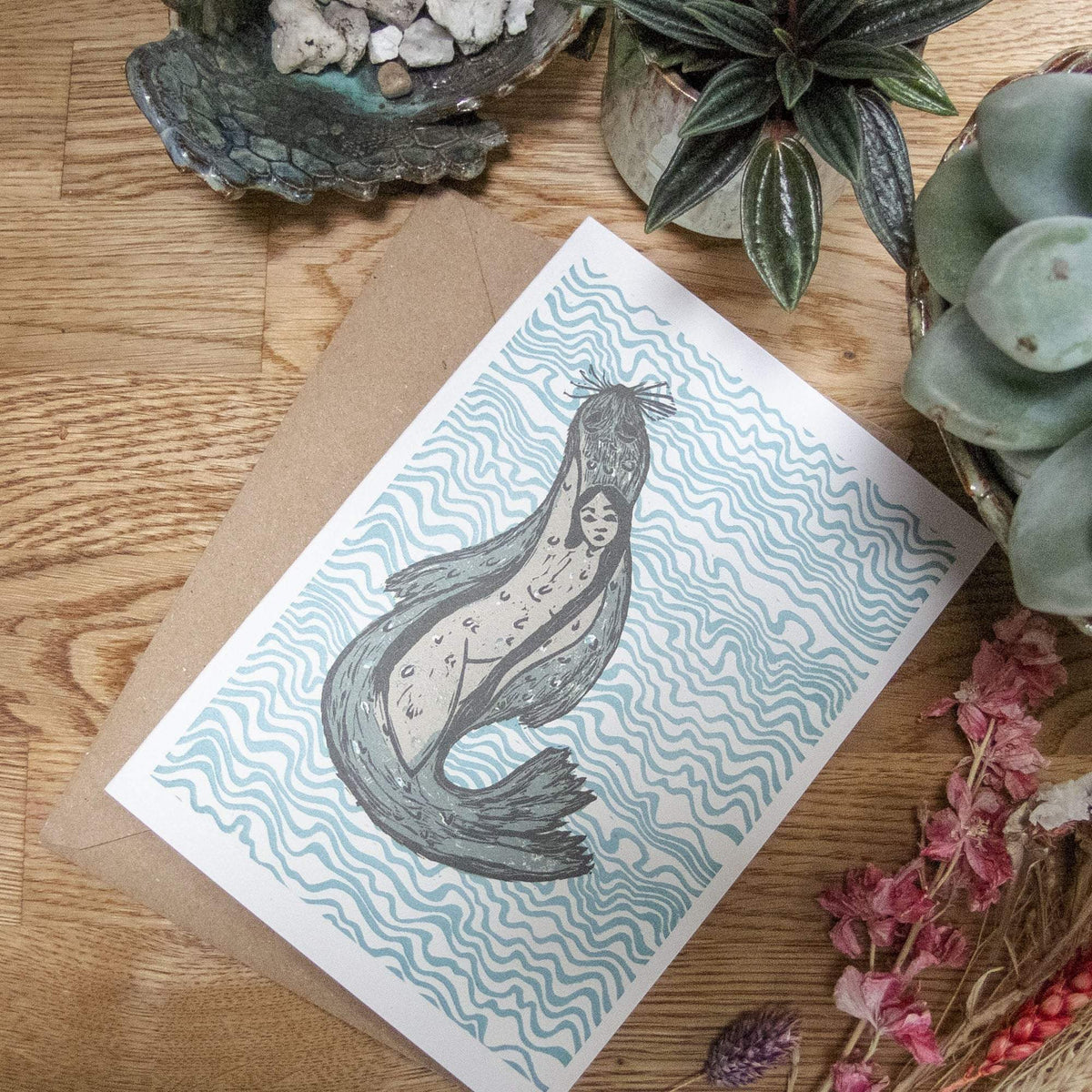 THE SELKIE Greeting card with blue and grey linocut of a seal woman amid wavy lines on white cardstock, with kraft envelope.